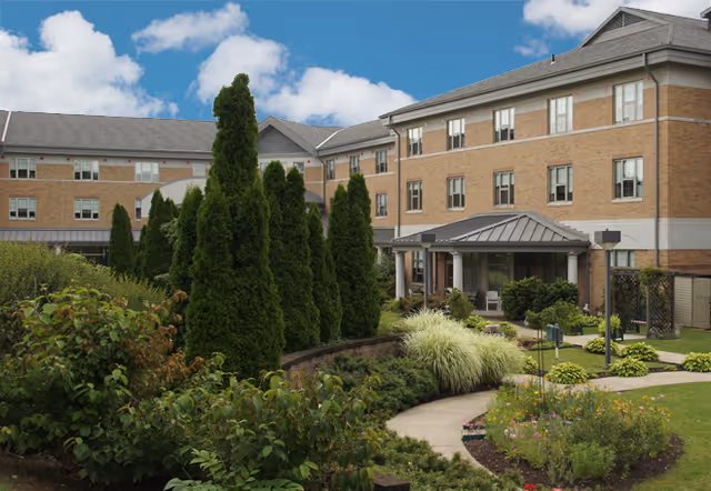 Exterior view of a multi-story brick building with a landscaped garden in the foreground, featuring tall evergreen trees, shrubs, and a curved walkway leading to a covered entrance. The sky is partly cloudy.