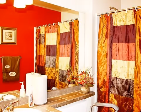 Bathroom with a colorful patchwork shower curtain in shades of orange, yellow, brown, and red. The bathroom has a red accent wall with a framed picture and brown towels hanging on a towel rack. A countertop with a sink, soap dispenser, paper towel holder, and a decorative plant is visible.
