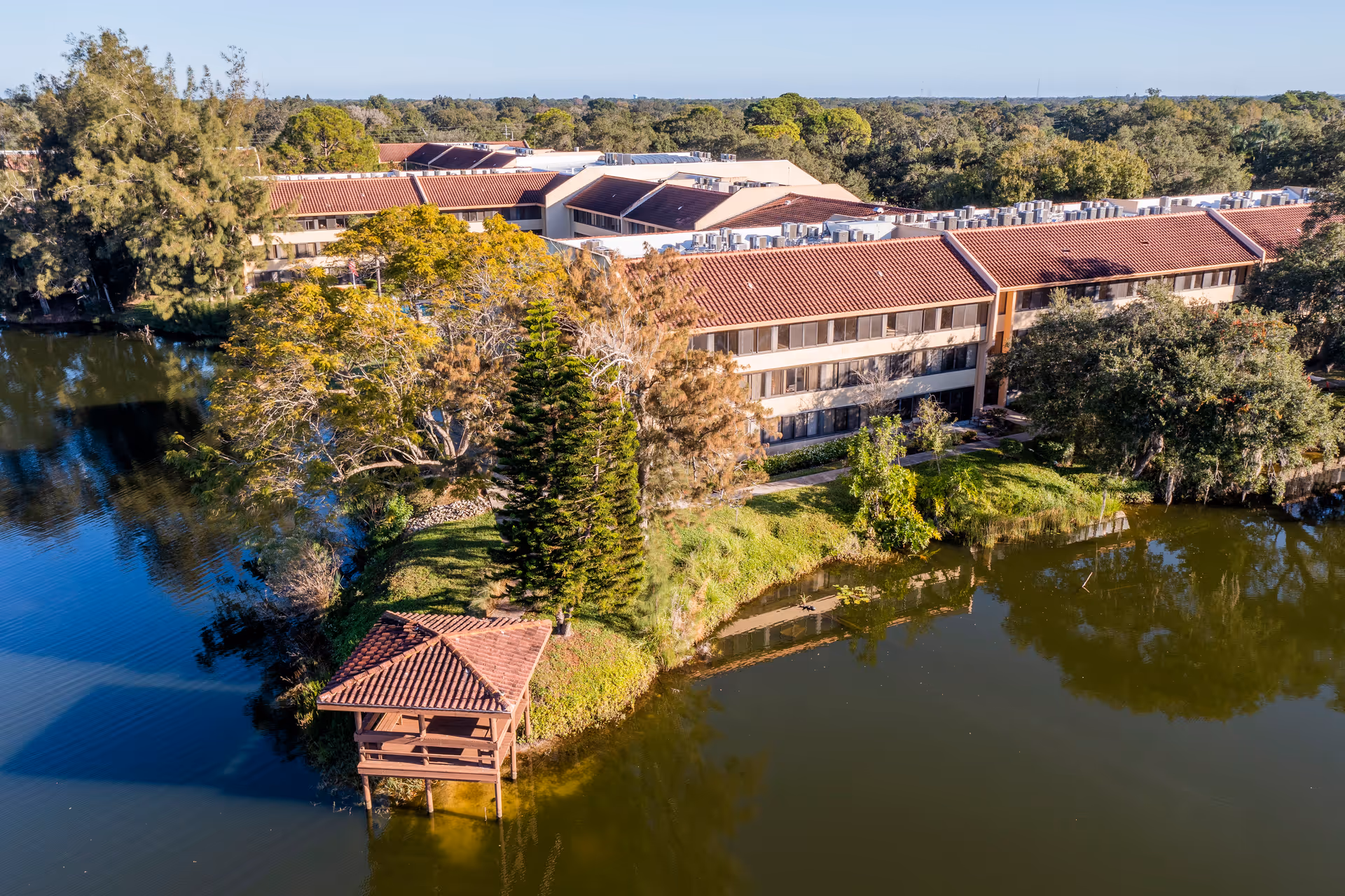 Aerial view of American House Sarasota, a senior living facility with a red-tiled roof, surrounded by trees and water. There is a small wooden gazebo on stilts over the water near the building.