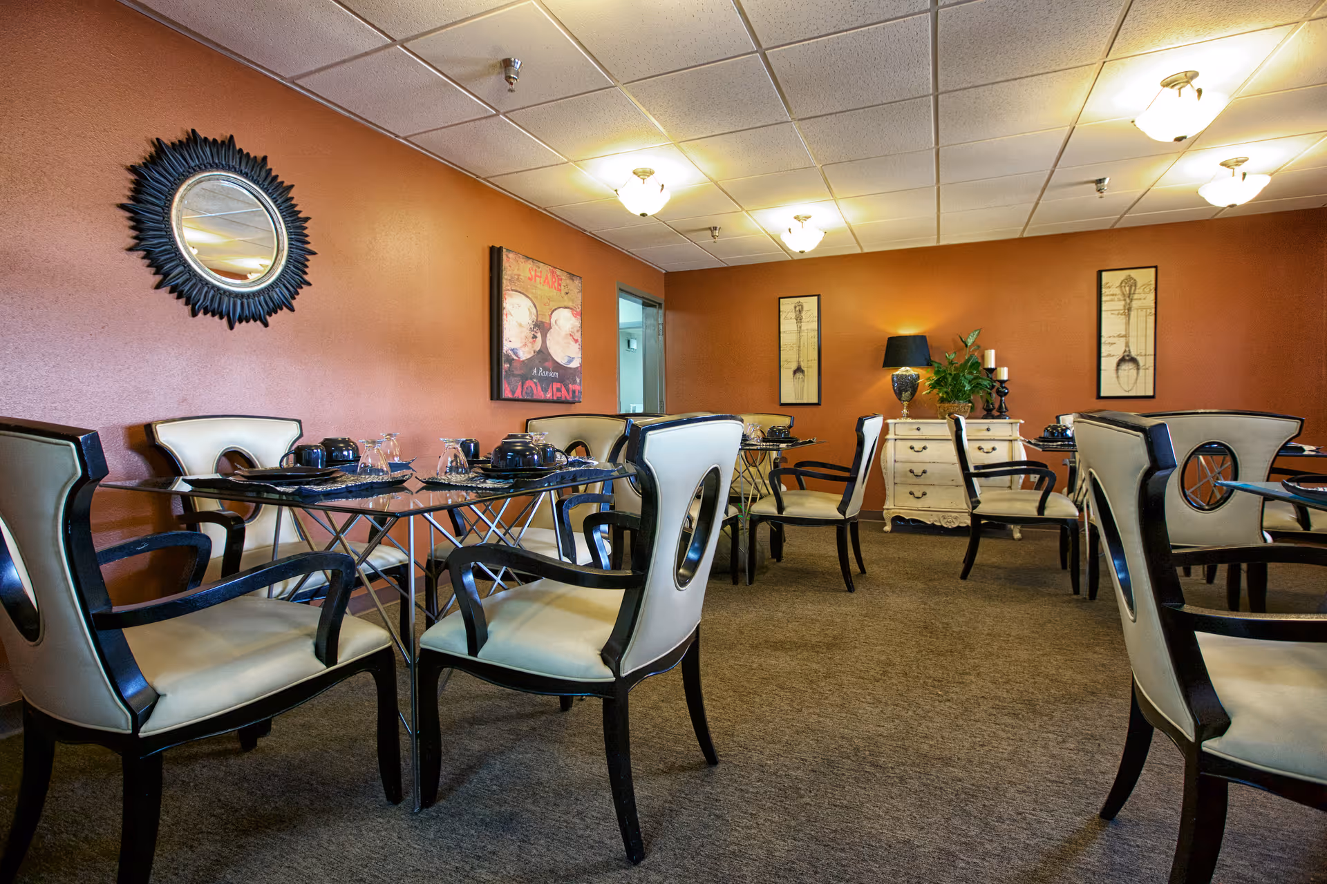 Dining room with several glass-top tables and cream-colored chairs with black frames. The walls are painted a warm orange color and decorated with framed artwork and a round mirror. A small white dresser with a lamp, plant, and candles is against the far wall.