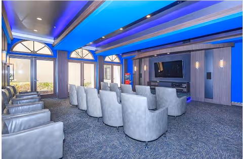 A modern media room with rows of gray upholstered swivel chairs facing a large flat-screen TV mounted on a wooden paneled wall. The room features blue accent lighting on the ceiling and carpeted flooring. Large windows with arched tops allow natural light to enter the space.