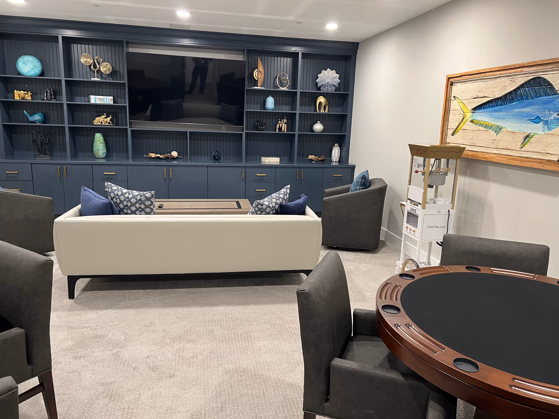 A cozy living room area featuring a white sofa with blue and patterned pillows, two gray armchairs, a round poker table with cup holders, and a large built-in dark blue shelving unit with decorative items and a flat-screen TV. A large framed painting of a fish hangs on the wall, and a popcorn machine is placed near the corner.