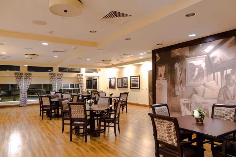 A spacious dining room with multiple dark wood tables and chairs arranged neatly on a polished wooden floor. The room is well-lit with ceiling lights and has large windows with patterned curtains. The walls feature framed black and white photographs and a large sepia-toned mural depicting a vintage scene with a man sitting on a bed and various items hanging on the wall.