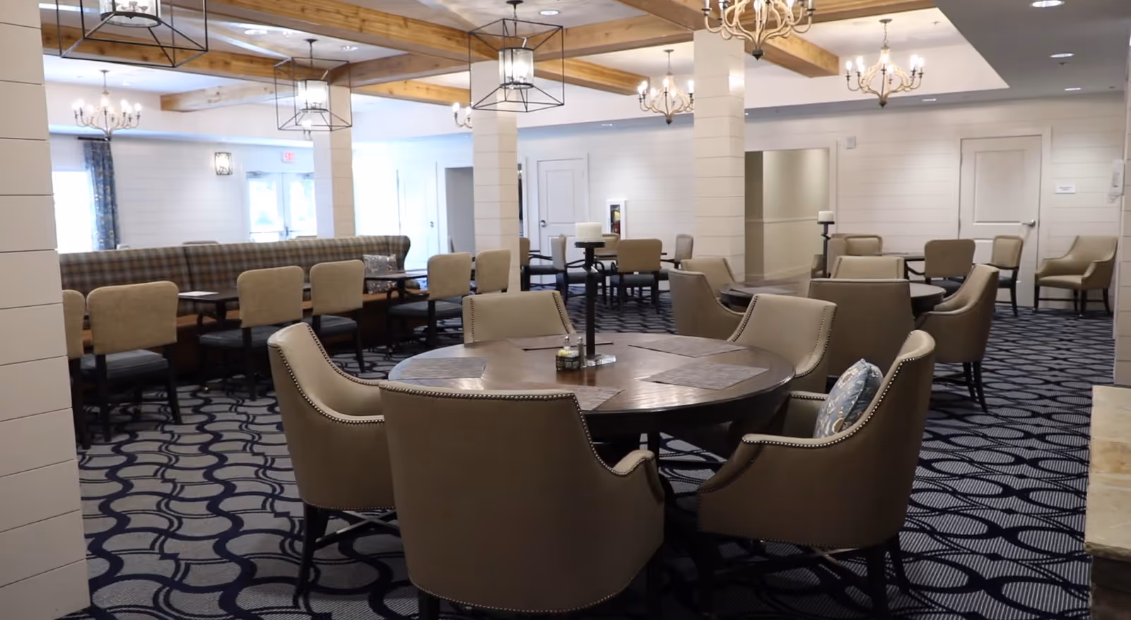 A spacious dining room with multiple tables and chairs arranged neatly. The room features a patterned carpet, wooden ceiling beams, and several chandeliers and pendant lights hanging from the ceiling. There are beige upholstered chairs around round and rectangular tables, with placemats and candle holders on the tables. The walls are light-colored with white paneling and doors visible in the background.