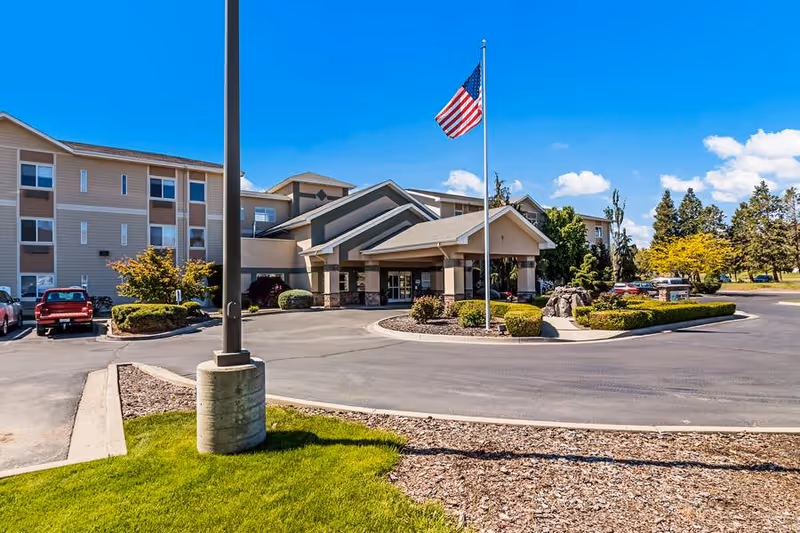 Exterior view of Rexford Park senior living facility with a circular driveway, landscaped bushes, an American flag on a flagpole, and a clear blue sky.