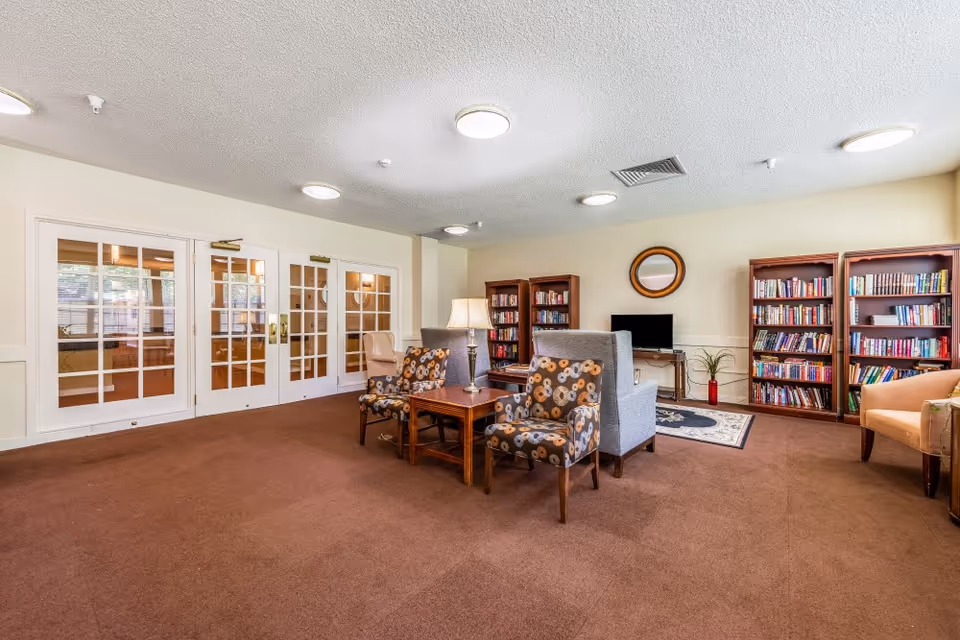 A cozy lobby area featuring two patterned chairs, a small wooden table, and bookshelves filled with books. Glass doors lead to another room, and a television is mounted on the wall.