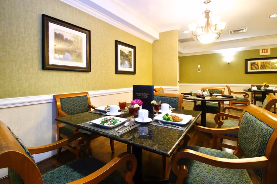Dining room with a set table holding plates, drinks and place settings surrounded by upholstered wooden chairs in a warmly lit senior living facility.