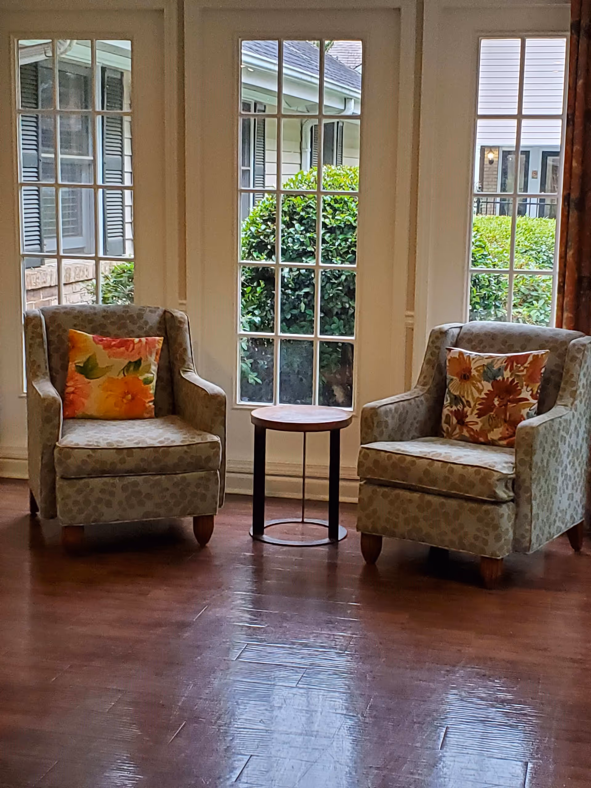 Two patterned armchairs with floral cushions are positioned on either side of a small round wooden table in front of large windows showing green bushes outside. The room has wooden flooring and white window frames.