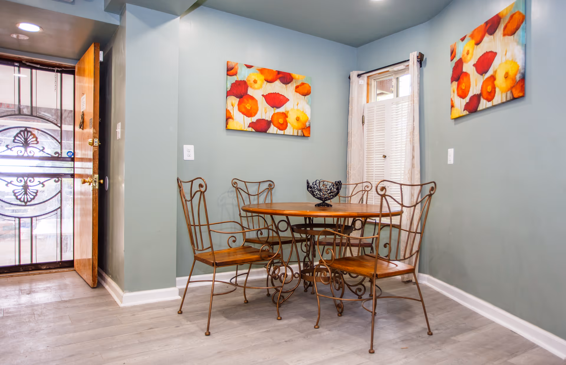 Small dining area with a round wooden table and four wrought-iron chairs against teal walls decorated with bright floral paintings and a door with a decorative metal screen.