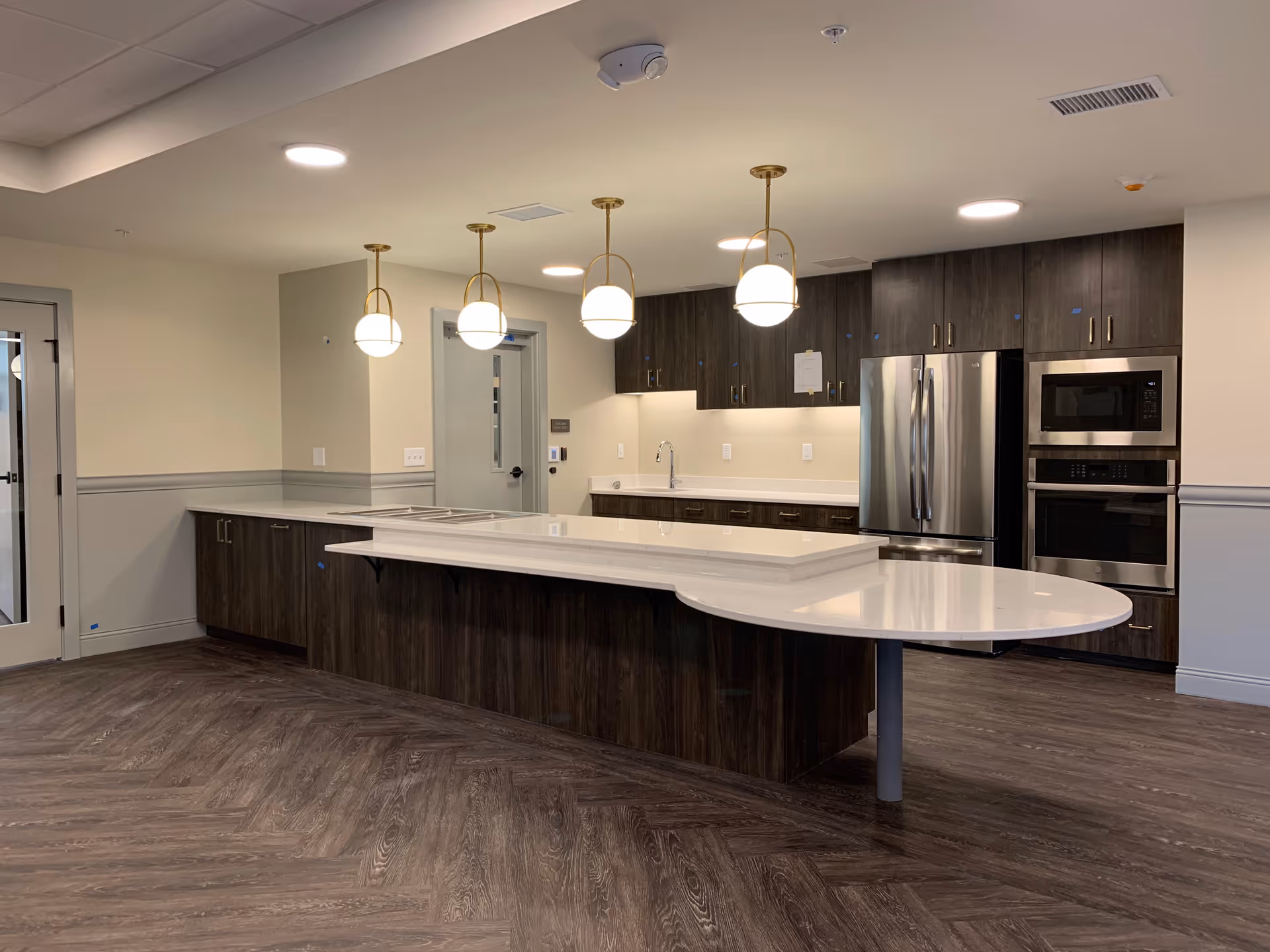 Modern kitchen area with dark wood cabinets, a large white countertop island with rounded edges, stainless steel refrigerator, built-in microwave and oven, and four pendant lights hanging from the ceiling. The floor has a dark wood pattern and the walls are painted light beige with gray wainscoting.