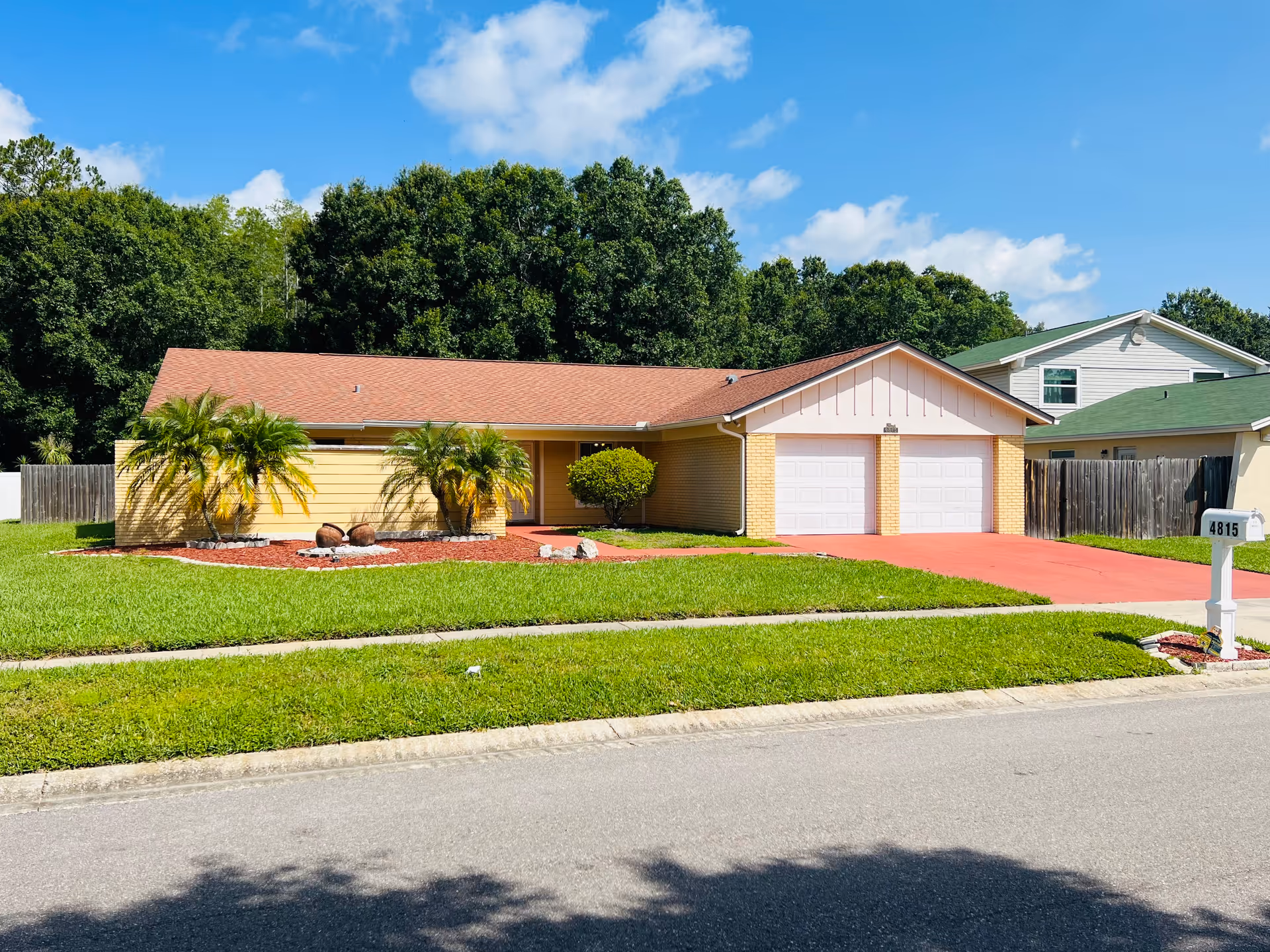 Single-story residential building with a red roof and yellow brick exterior, featuring a double garage with white doors. The front yard has green grass, palm trees, and a red driveway. A white mailbox with the number 4815 is visible near the street. Trees and a blue sky with some clouds are in the background.