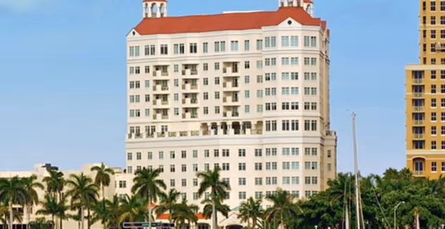 Exterior view of a multi-story residential building with a red roof, surrounded by palm trees and other greenery under a clear blue sky.
