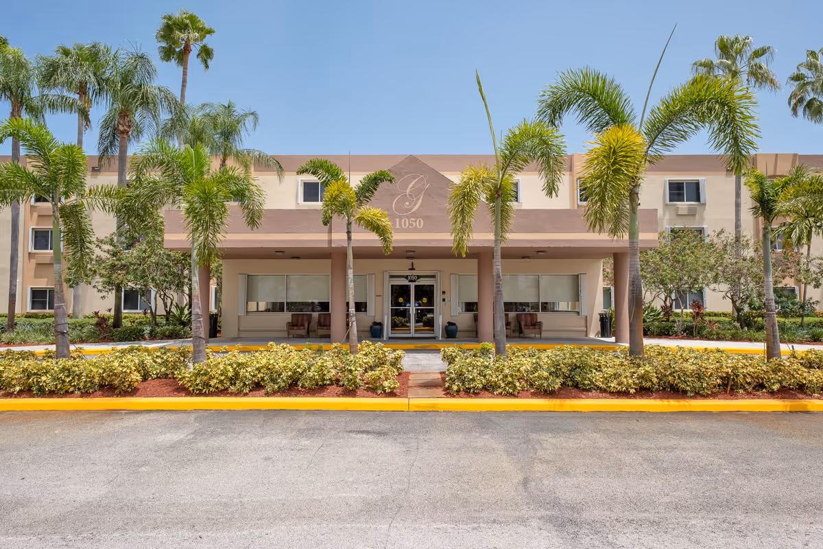 Front exterior view of the Grand Villa of Deerfield Beach building with palm trees and landscaped bushes in front under a clear blue sky.