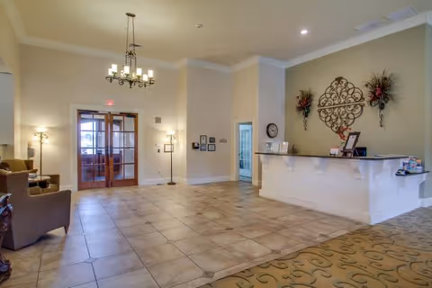 A spacious and well-lit reception area in a senior living facility with a tiled floor, a white reception desk adorned with decorative wall art and floral arrangements, a chandelier hanging from the ceiling, and seating area with armchairs near the entrance with glass double doors.