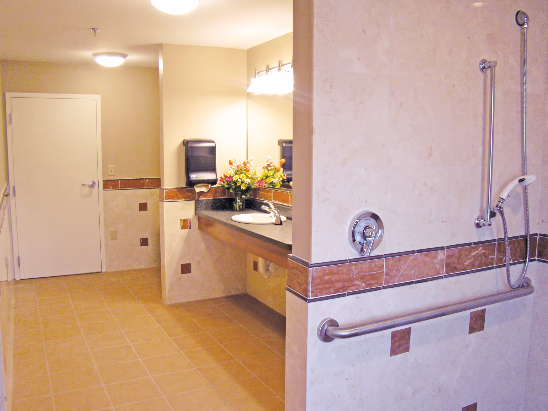 A clean, spacious bathroom with beige tiled floors and walls. The bathroom features a white door, a large mirror above a black countertop with a sink, a paper towel dispenser, and a vase with colorful flowers. There is a shower area with a handheld showerhead, grab bars, and a control knob on the wall.