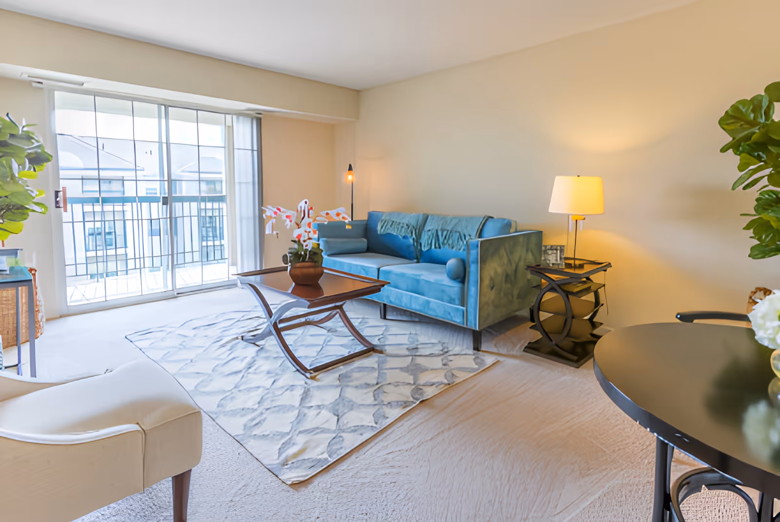 A bright living room with a blue sofa, a wooden coffee table with a potted plant, a beige armchair, a side table with a lamp, and large sliding glass doors leading to a balcony.