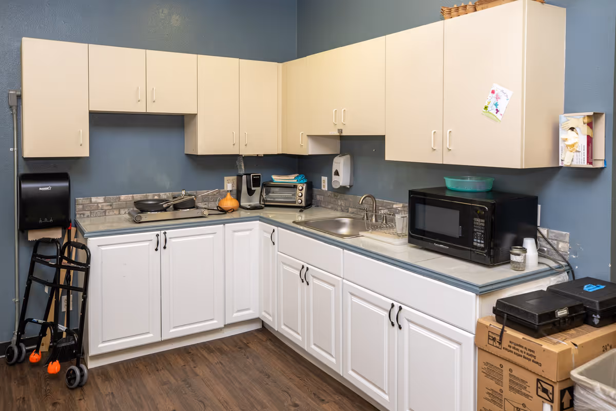 A small kitchen area with white lower cabinets and beige upper cabinets against blue walls. The countertop holds a microwave, toaster oven, coffee maker, double hot plate with pans, a sink, and some small containers. A black paper towel dispenser and a hand truck are positioned in the corner. There are cardboard boxes and a trash bin on the right side.