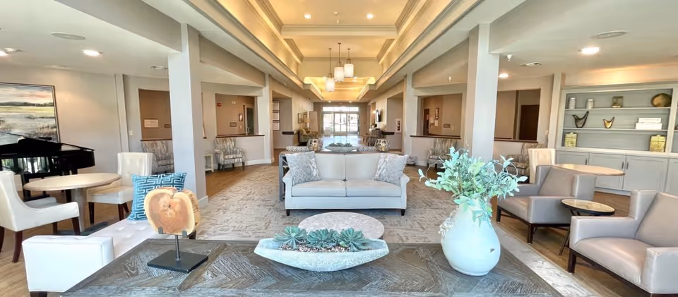 A spacious and well-lit senior living facility common area with comfortable seating including sofas and armchairs arranged around small tables. The room features neutral tones, decorative plants, artwork on the walls, and a grand piano on the left side. The ceiling has recessed lighting and pendant lights, and there is a large window or glass door at the far end letting in natural light.