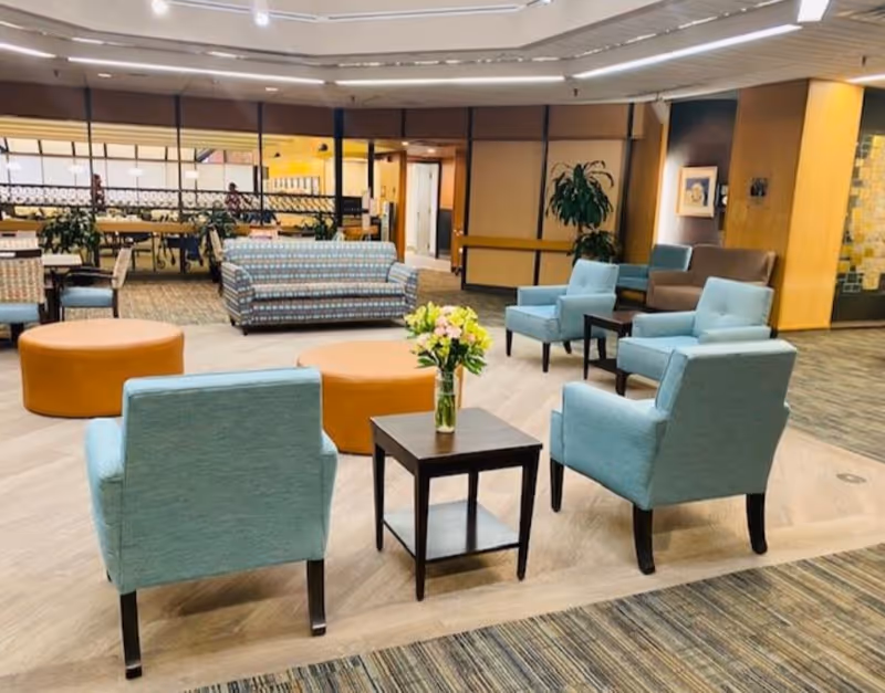 A spacious senior living facility common area with light blue armchairs arranged around a small dark wooden table holding a vase of flowers. In the background, there are patterned sofas, round orange ottomans, plants, and a dining area separated by glass partitions.