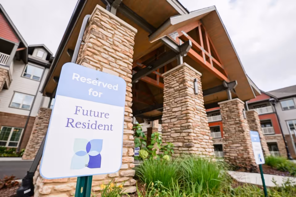 Entrance of a senior living facility with stone pillars and a wooden roof structure. A sign in the foreground reads 'Reserved for Future Resident'. The building has multiple windows and balconies, with greenery and plants around the entrance.