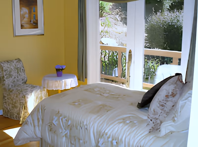 Sunlit bedroom with a made bed, sliding glass doors to a balcony, a small round table with a purple flower pot and an upholstered chair.