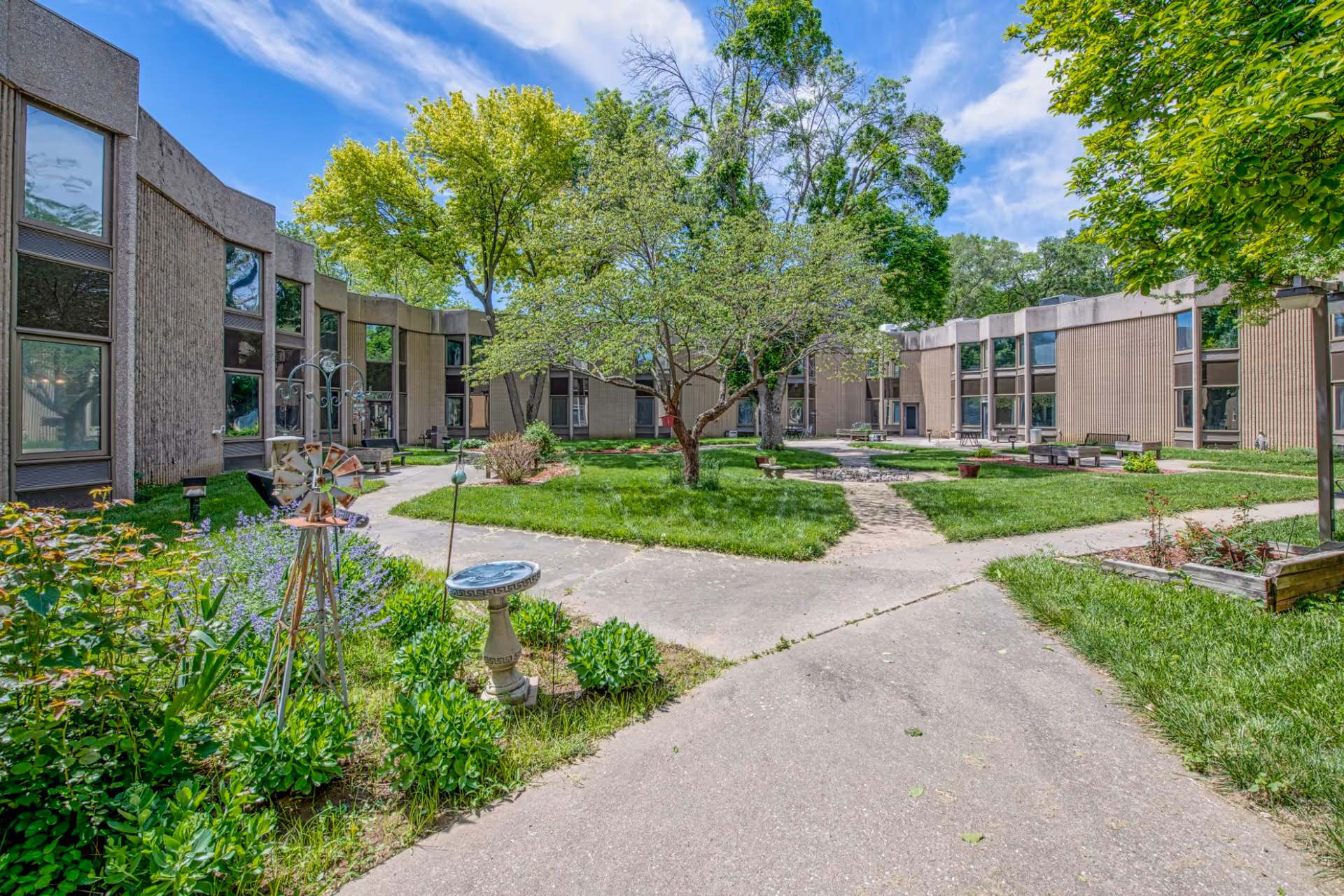 Outdoor courtyard area at Bethany Lutheran Health Services featuring a paved walkway, green grass, trees, garden plants, and benches surrounded by a two-story building under a blue sky with some clouds.