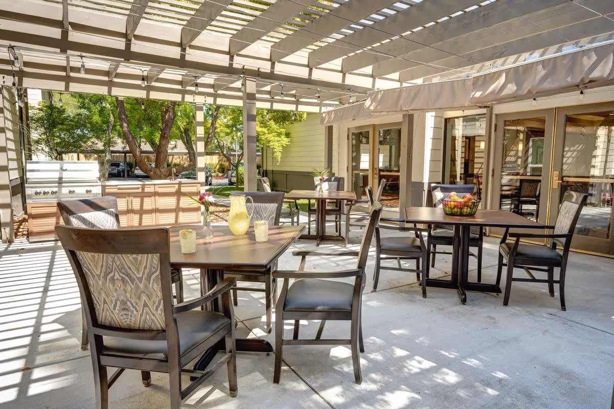 Outdoor patio area with several tables and chairs under a pergola. There is a grill station in the background and trees providing greenery. The patio is adjacent to a building with large glass doors.