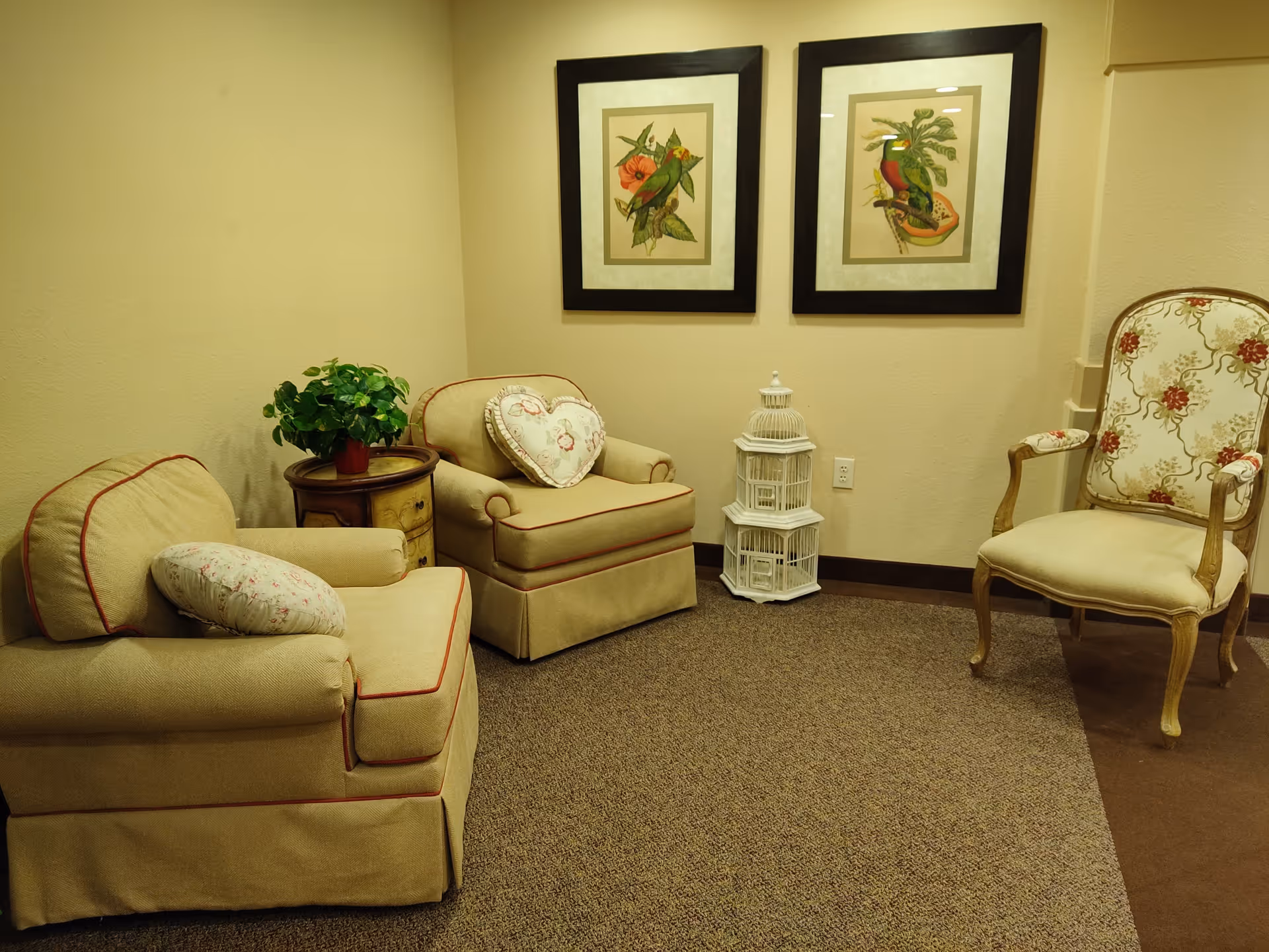 A cozy sitting area with two beige armchairs, one with a heart-shaped pillow and the other with a floral pillow, a small wooden side table with a green potted plant, a decorative white birdcage, and a floral upholstered wooden chair. Two framed bird illustrations hang on the beige wall behind the chairs.