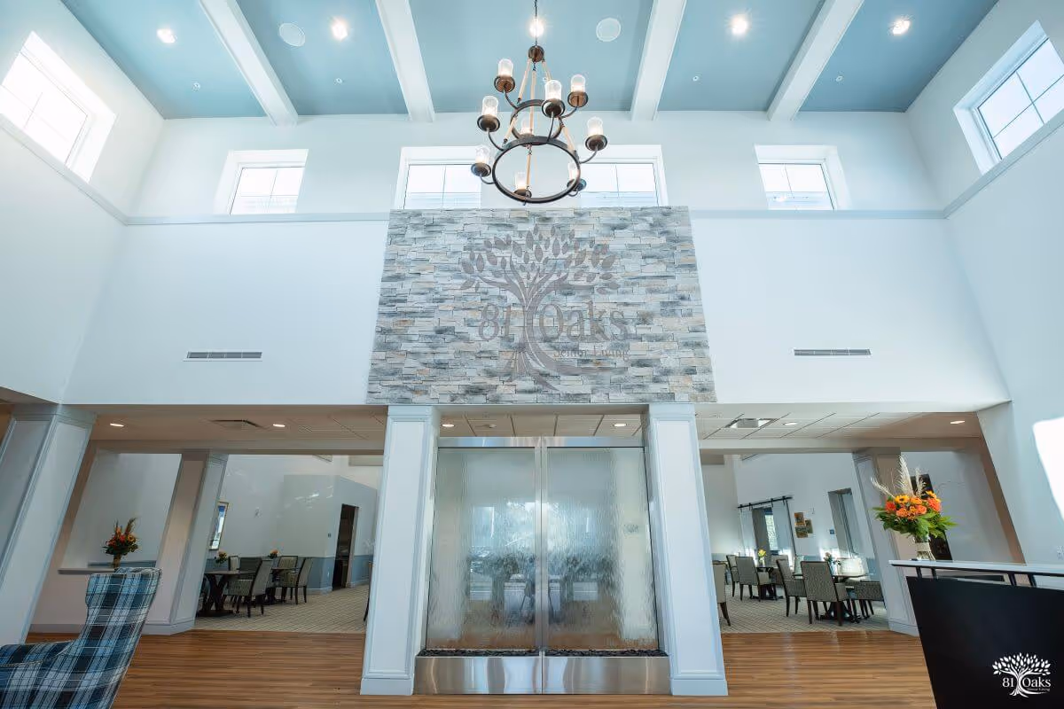 Bright and spacious senior living facility lobby with high ceilings, a chandelier, large windows, and a stone wall featuring the 81 Oaks logo. There are seating areas with chairs and tables on either side, and a water feature beneath the stone wall.