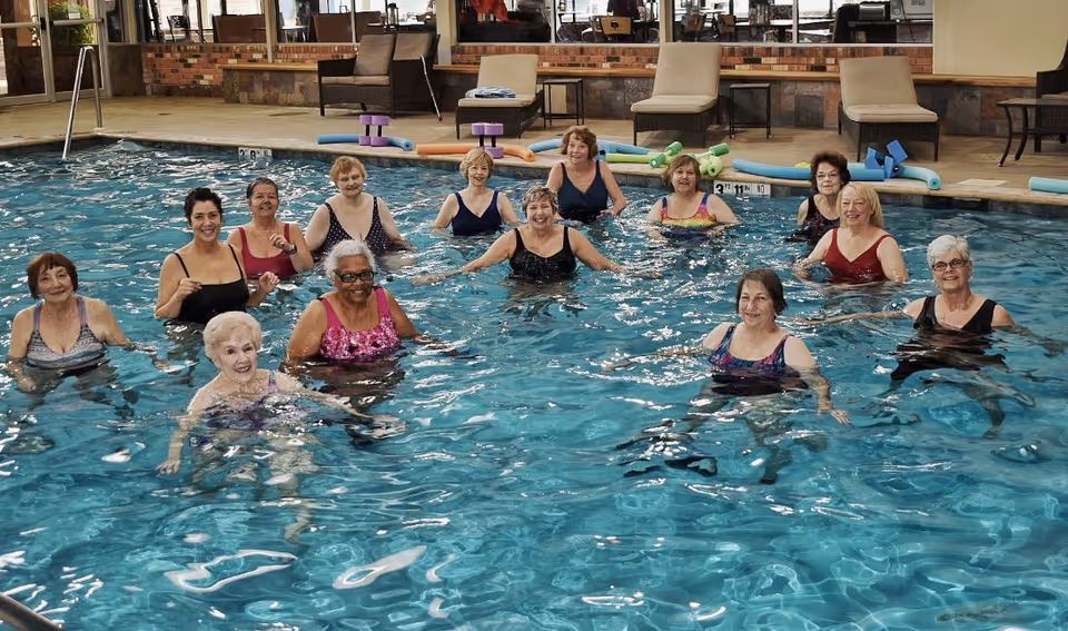 A group of elderly women standing and smiling in an indoor swimming pool, participating in a water exercise class. The pool area has lounge chairs and pool noodles along the edge.