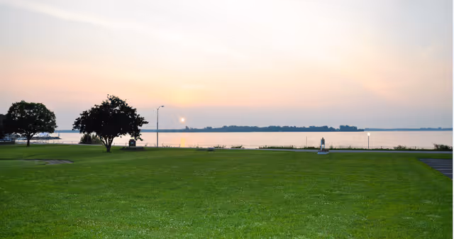 A grassy lawn with a few trees near the water's edge, overlooking a calm body of water at sunset with a clear sky.
