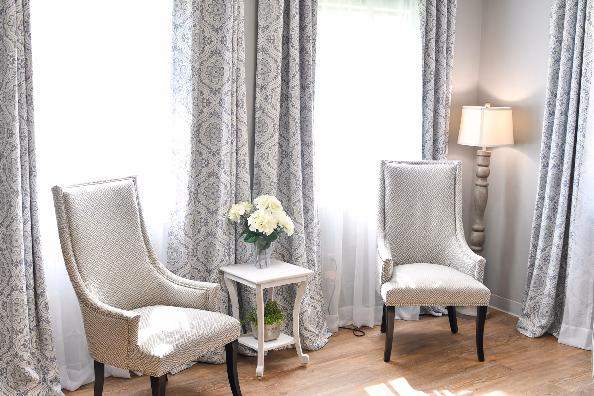 A bright sitting area with two patterned upholstered chairs facing each other, separated by a small white side table holding a vase of white flowers. The background features large windows with sheer white curtains and decorative patterned drapes. A floor lamp with a beige shade stands in the corner on a wooden floor.