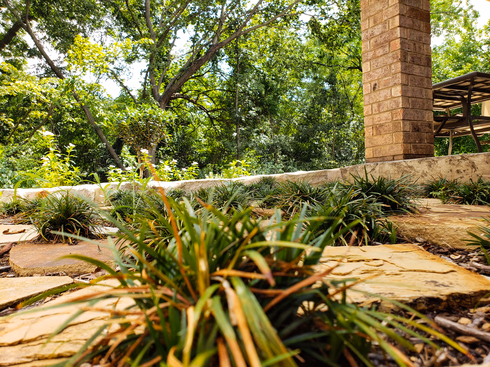 Low-angle view of a landscaped outdoor patio with stone pavers, low ornamental grasses, a brick column and trees in the background.