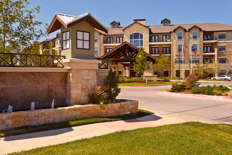 Exterior view of Waterview The Point Independent Living facility showing a multi-story building with stone and beige walls, a covered entrance with wooden beams, landscaped greenery, a small water feature with fountains, and flags near the driveway.