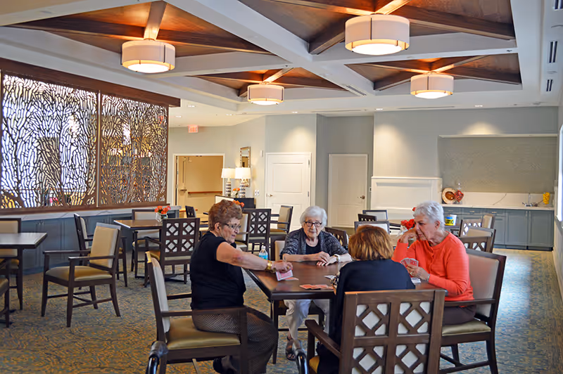 Four older adults sit around a table playing cards in a bright, spacious common dining area with decorative ceiling lights and a patterned room divider.