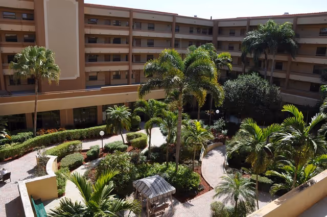 View of a landscaped courtyard garden with palm trees, shrubs, and a small wooden gazebo, surrounded by a multi-story residential building with balconies.