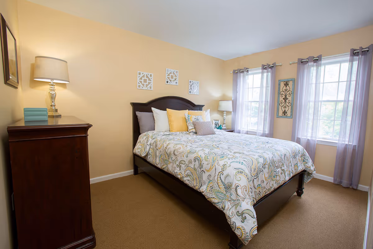 A cozy bedroom with a large bed featuring a patterned comforter and multiple pillows. The room has beige walls, two windows with sheer lavender curtains, a wooden dresser with a lamp and decorative items, and wall art above the bed.