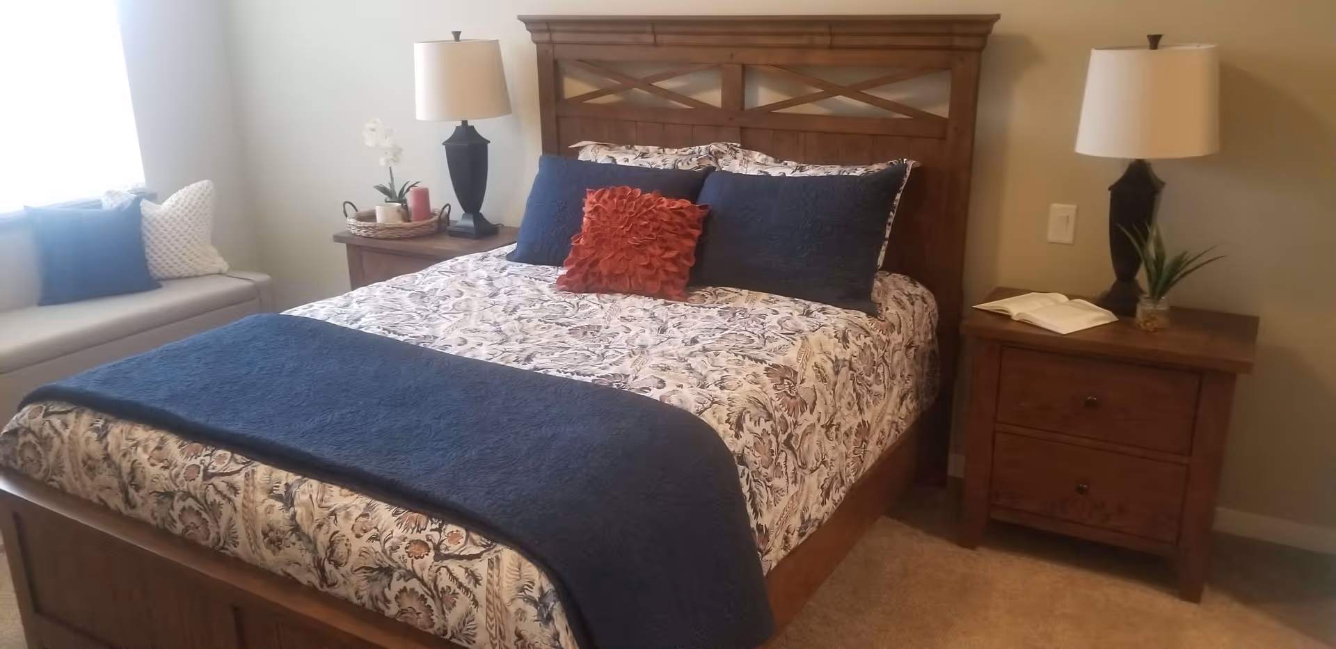 A cozy bedroom with a wooden bed frame and headboard, a bed made with patterned bedding and blue and red decorative pillows. There are two wooden nightstands on either side of the bed, each with a lamp. One nightstand has a small plant and an open book, while the other has a tray with candles and a white orchid. A cushioned bench with pillows is positioned near a window with natural light coming through.