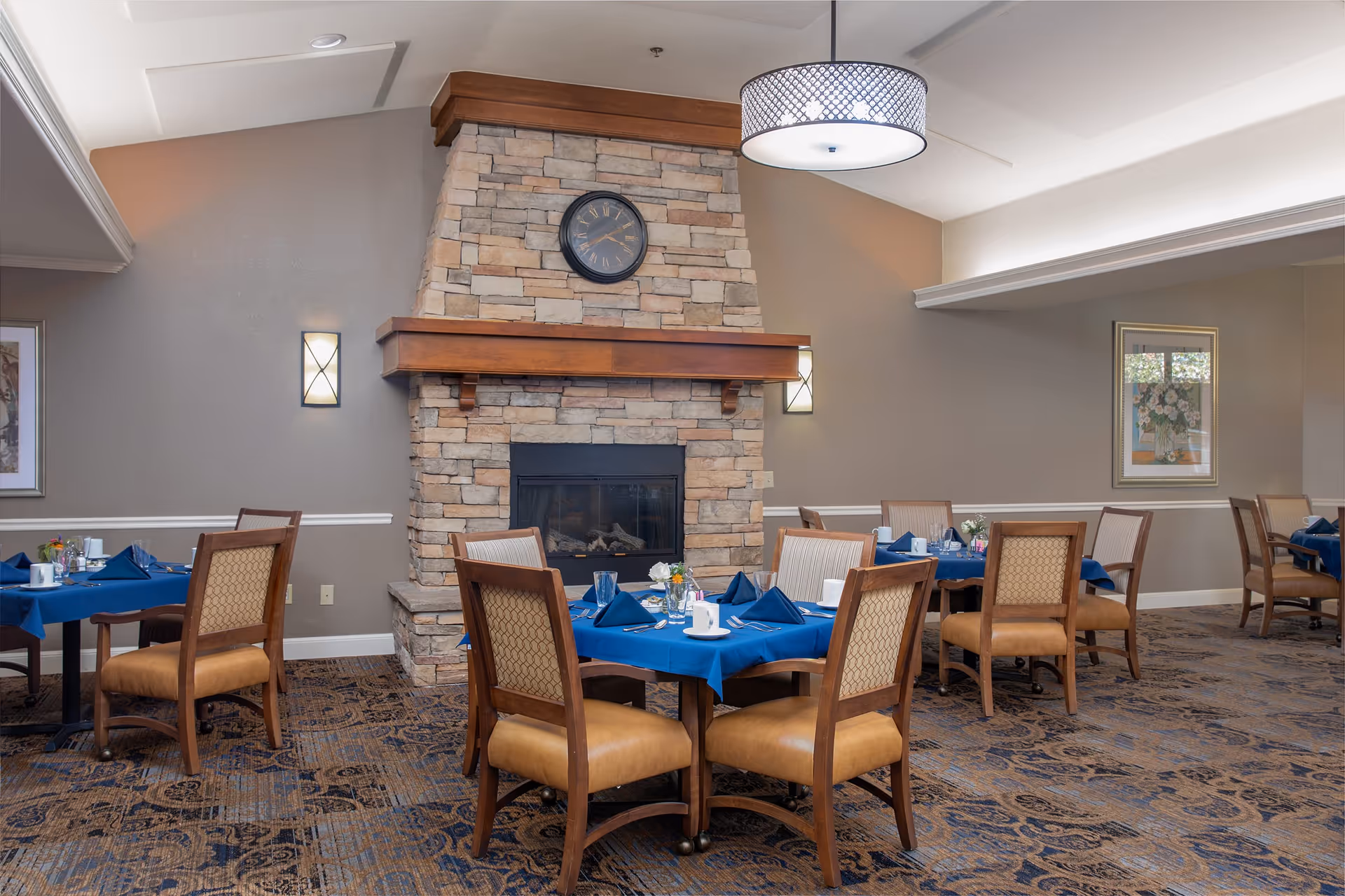 Dining room with several wooden tables covered with blue tablecloths, each set with napkins, cups, and silverware. The room features a stone fireplace with a wooden mantle and a clock above it. There are upholstered chairs around the tables and framed artwork on the walls.