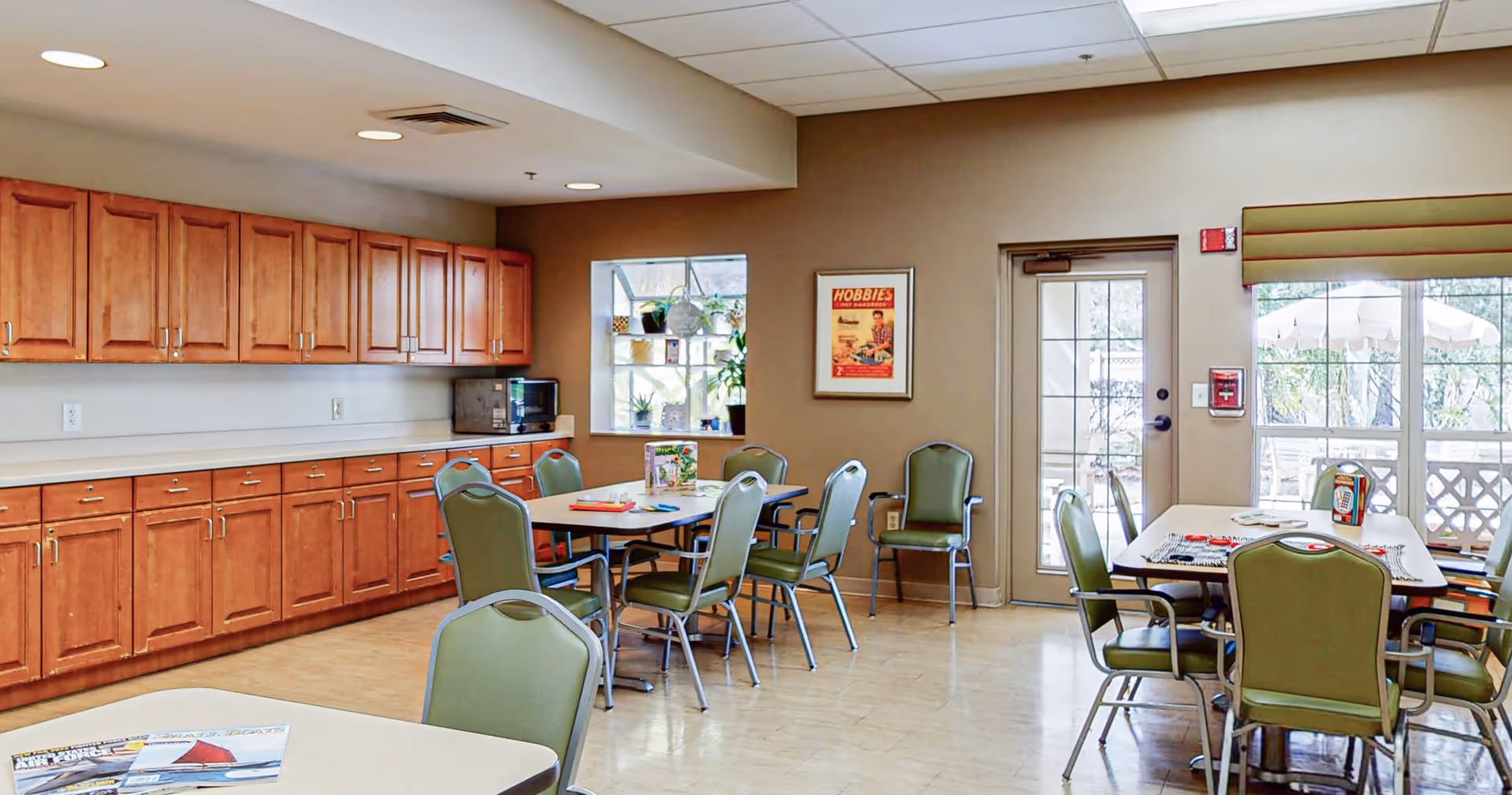 A well-lit room with multiple tables and green chairs arranged around them. The room features wooden cabinets along one wall, a microwave on the countertop, a window with plants on the sill, a framed poster on the wall, and a glass door leading outside. The floor is light-colored, and the ceiling has recessed lighting.