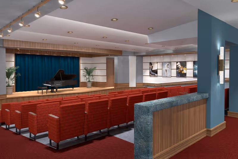 An auditorium with rows of red cushioned seats facing a wooden stage with a black grand piano and two potted plants. The back wall of the stage has a dark blue curtain. The room features recessed ceiling lights, framed pictures on the side wall, and a blue column with a wall sconce light fixture.