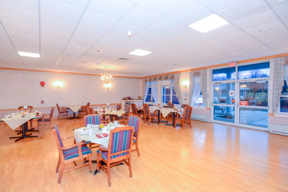 A bright dining room with several tables set with cups, plates, and napkins. The room has wooden floors, patterned wallpaper, and large windows with curtains. There are wooden chairs with colorful striped cushions around the tables. A chandelier and wall sconces provide lighting, and a glass door leads outside.
