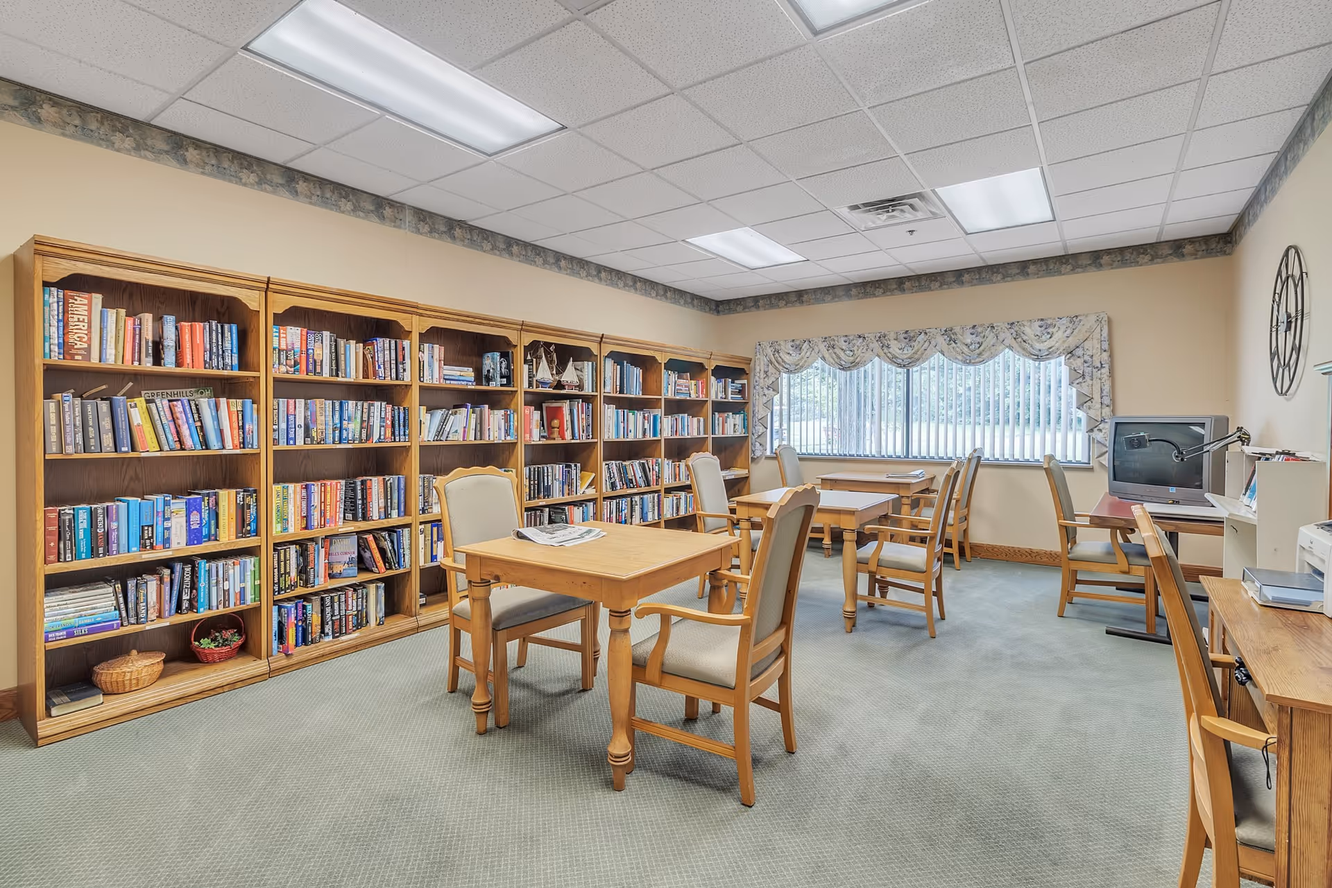 A quiet reading room with multiple wooden bookshelves filled with books along one wall. Several wooden tables with cushioned chairs are arranged throughout the room. A large window with vertical blinds and floral valance lets in natural light. A computer workstation is set up in the corner with a monitor and desk lamp. The room has a carpeted floor and a drop ceiling with fluorescent lighting.
