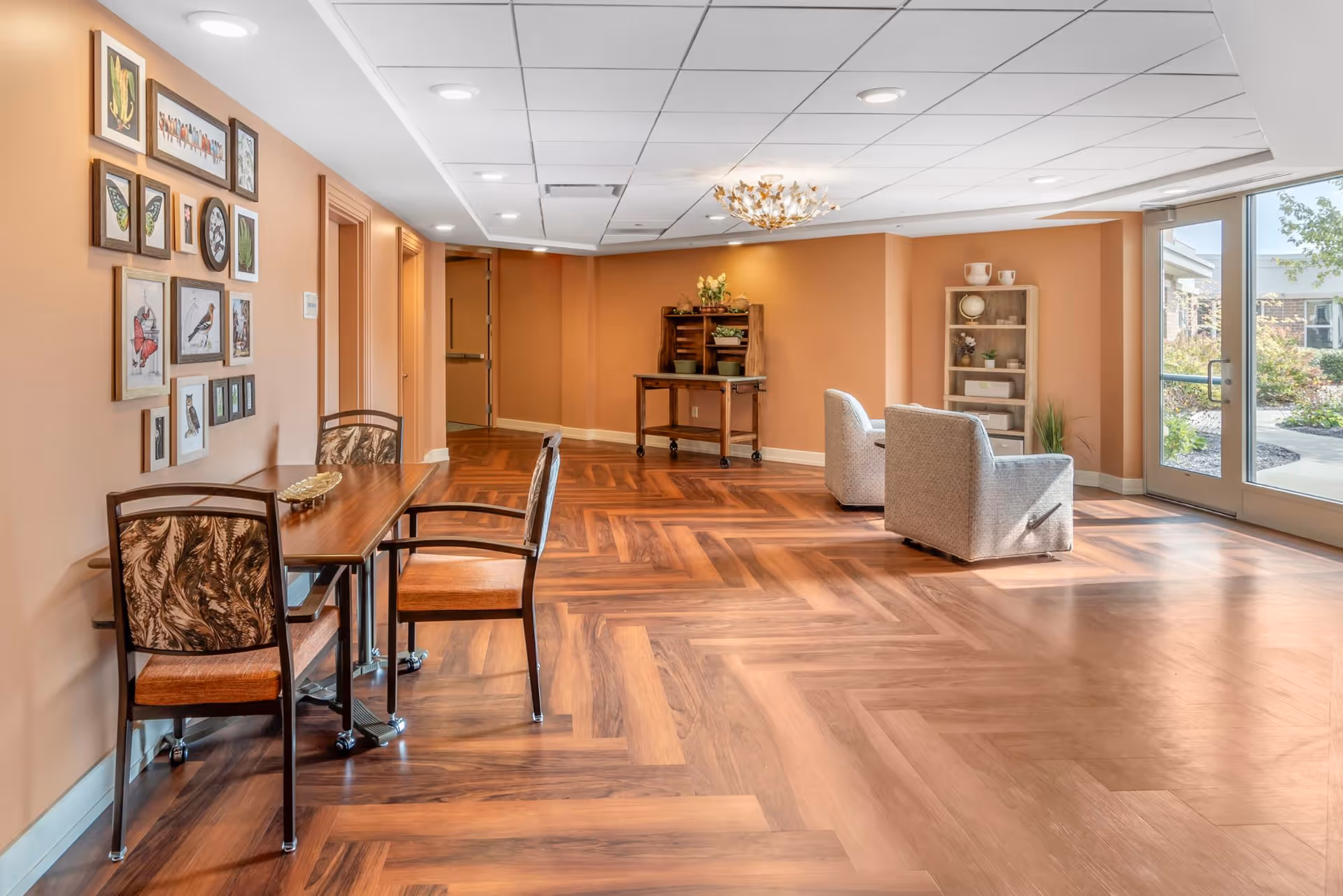 A bright and spacious common area with wood-patterned flooring and peach-colored walls. There is a small table with two chairs on the left side, a wall decorated with framed pictures of birds and butterflies, two cushioned armchairs near a large glass door leading outside, and a wooden shelving unit with decorative items. The ceiling has recessed lighting and a decorative chandelier.