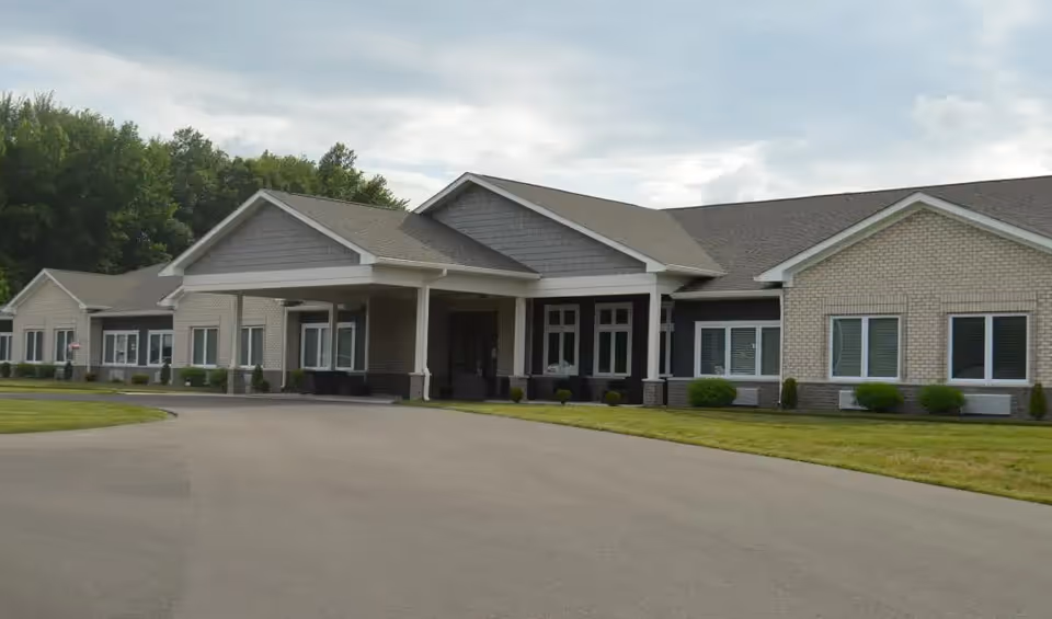 Exterior view of a single-story senior living facility building with a covered entrance, surrounded by a paved driveway and green lawn, under a cloudy sky.