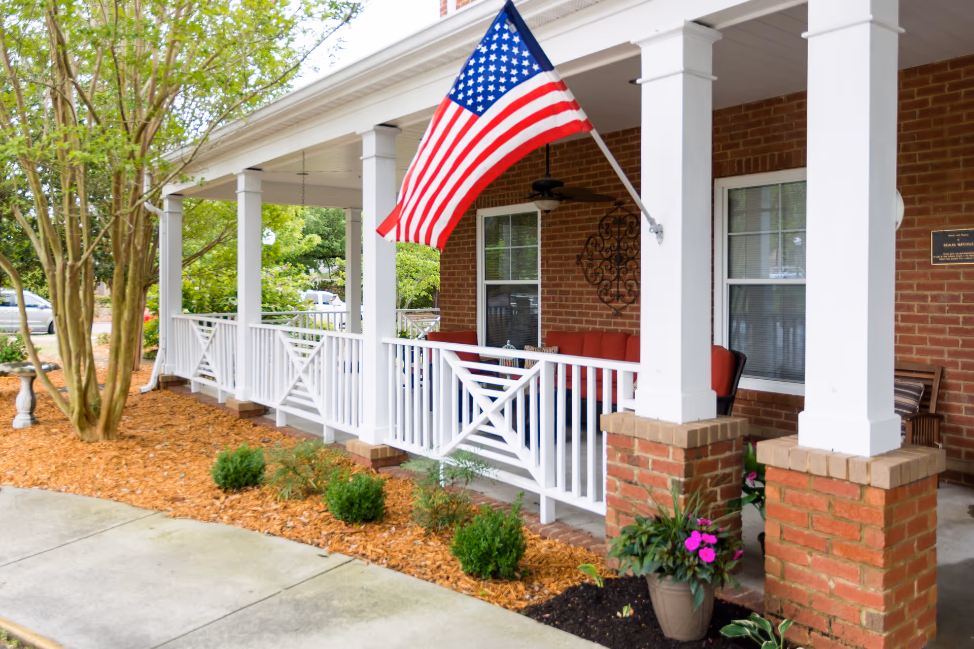 Front porch of a brick building with white columns and railing, an American flag mounted on one of the columns, outdoor seating with red cushions, potted plants, and a tree nearby.