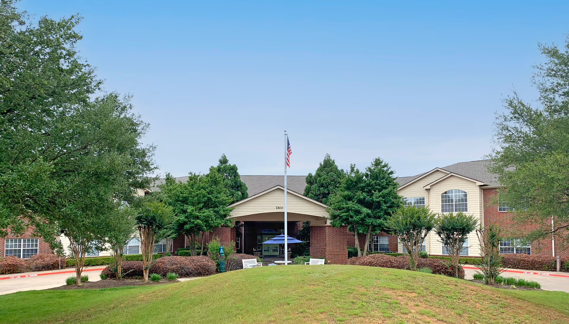 Front exterior view of Morada Lake Arlington facility showing a two-story brick building with beige siding, surrounded by green trees and shrubs, with a flagpole and benches in front on a grassy area under a clear blue sky.