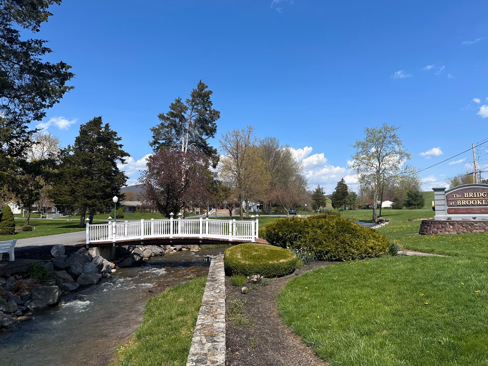 A small white pedestrian bridge over a flowing creek surrounded by green grass, bushes, and trees under a clear blue sky. A sign on the right reads 'The Bridge at Brookline'.