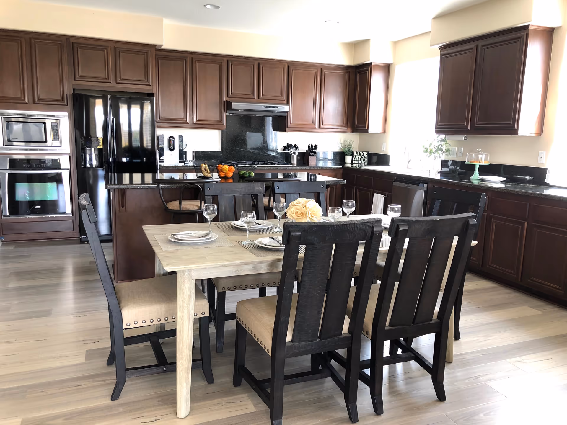 A modern kitchen and dining area with dark wooden cabinets, black granite countertops, and stainless steel appliances including a refrigerator, oven, and microwave. A light wood dining table is set with plates, glasses, and a centerpiece of white roses, surrounded by six dark wooden chairs with beige cushions. The room has light-colored wooden flooring and large windows letting in natural light.