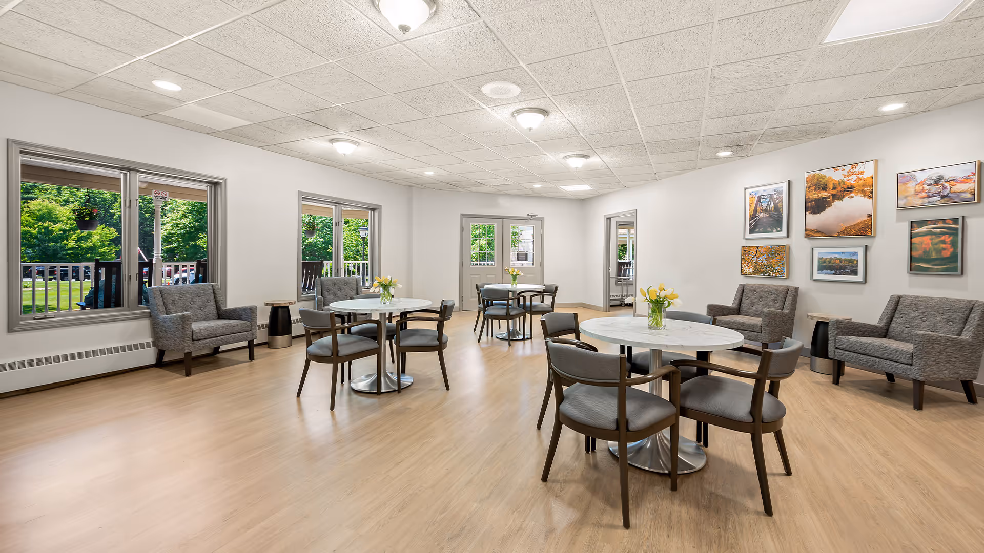 A bright and spacious common area with light wood flooring and white walls. The room features several round tables with four chairs each, decorated with small vases of yellow flowers. There are also gray upholstered armchairs and loveseats along the walls. Large windows and a glass door provide a view of green outdoor trees and a deck area. Framed landscape photographs hang on one wall.